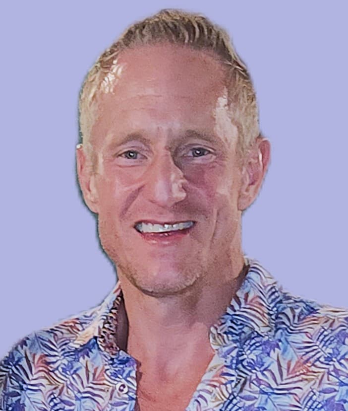 Smiling man with short blond hair wearing a blue and white tropical print button-down shirt.